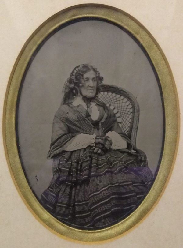Photo of Elizabeth Glasson in an oval frame. She is sitting very upright in a wicker chair, wearing heavy Victorian clothes: a layered dress with horizontal stripes a white broderie anglaise blouse and black lace gloves and headdress. Her hair is curled and styled into ringlets around her face., 
